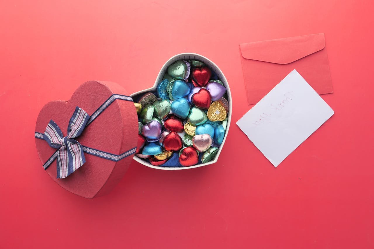 Heart-shaped box with colorful chocolates and envelope on a pink surface, perfect for Valentines.